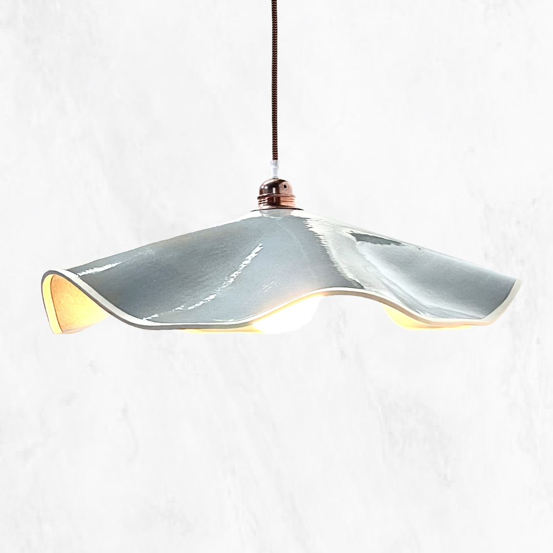 Eucalyptus Marilyn handmade ceramic pendant light with a flowing sculptural ceramic shade, glossy eucalyptus glaze, warm illuminated interior and copper suspension, shown against a light textured background.