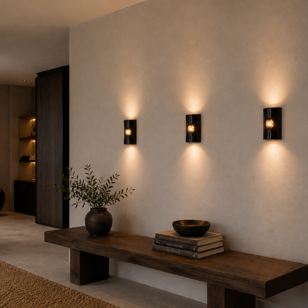 Three handcrafted ceramic wall sconces in a glossy graphite-black finish with illuminated amber textured glass inserts in a warm minimalist contemporary hallway with dark timber furniture and textured plaster walls.