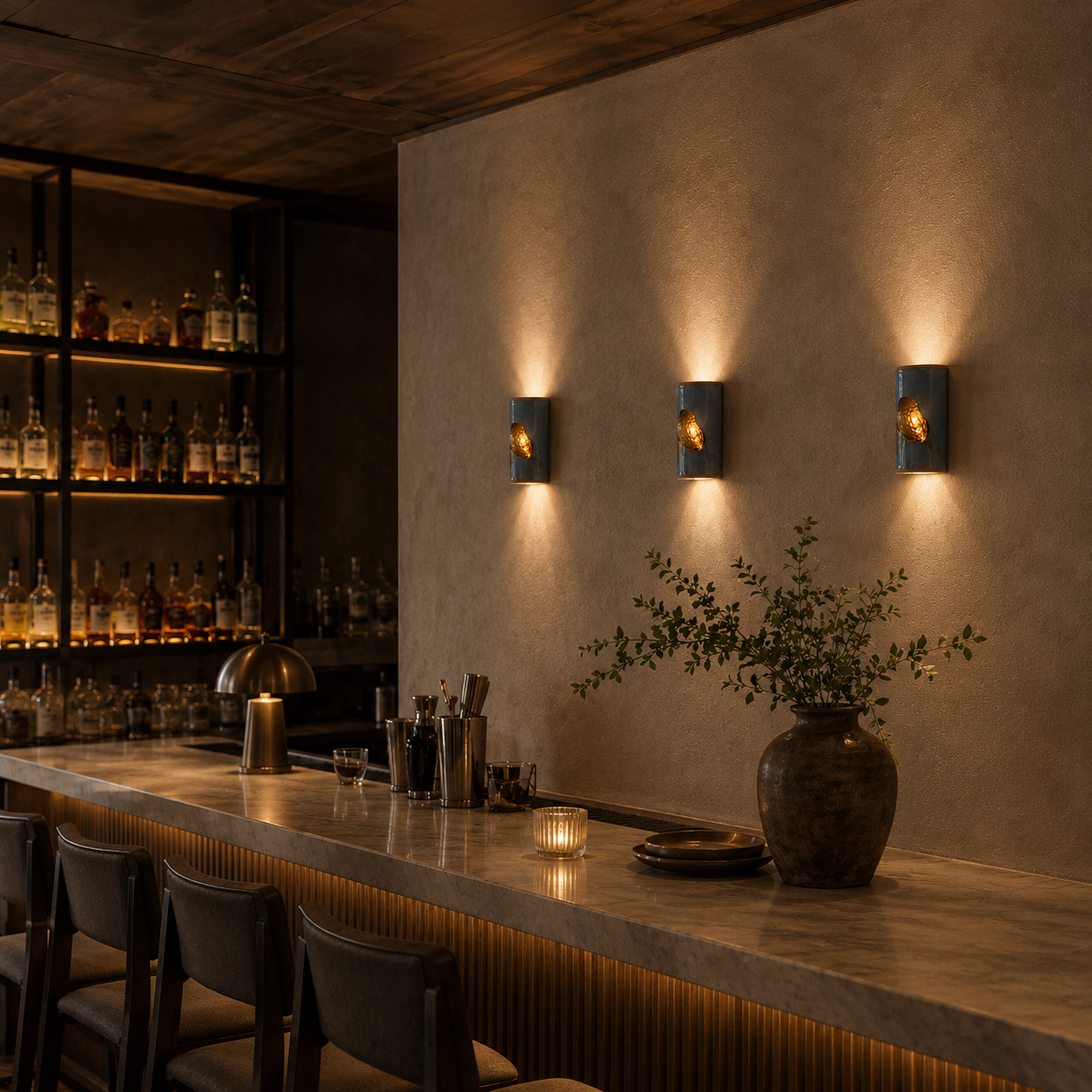 Three handcrafted ceramic wall sconces in a glossy midnight-blue finish with illuminated amber textured glass inserts in a moody contemporary bar interior with stone surfaces, timber detailing and warm ambient lighting.