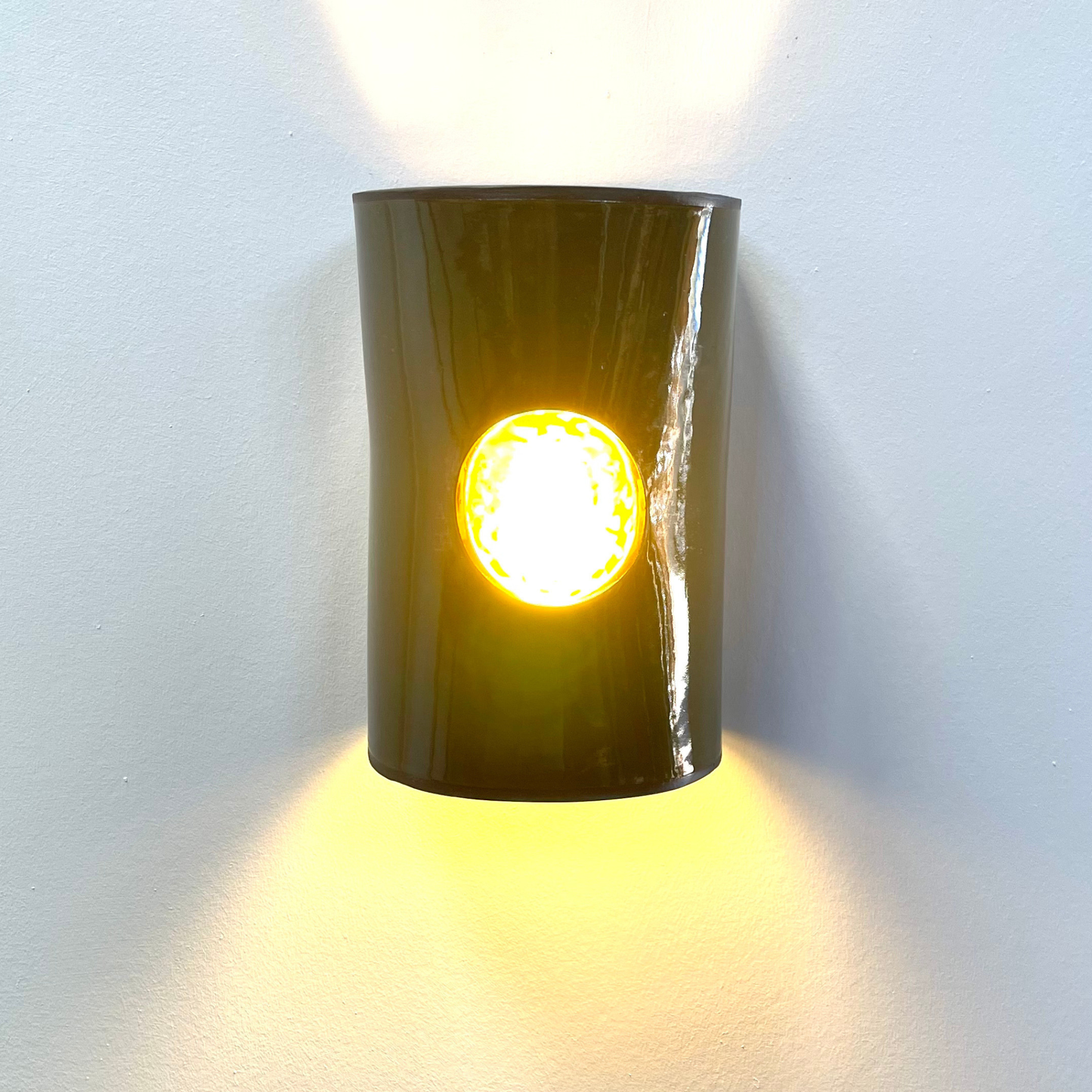 Handcrafted olive ceramic wall light with a glowing amber glass insert, casting warm up and down light against a textured wall.